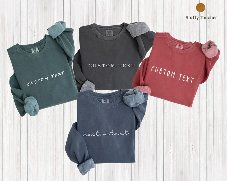 May include: Four long-sleeved sweatshirts in teal, charcoal, red, and navy blue, each with "CUSTOM TEXT" printed in white. The cuffs are folded up, revealing a lighter fabric. The sweatshirts are displayed on a light wood-grain background.