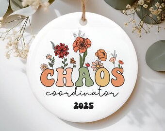 May include: A white ceramic ornament featuring a floral design and the words "CHAOS coordinator 2025". The text is in a retro font with orange, green, and yellow letters. The ornament has a string for hanging.