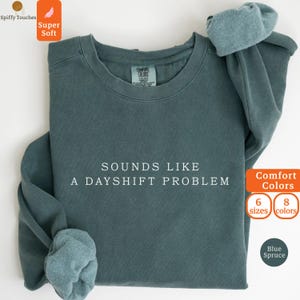 Può includere: Una felpa girocollo color blu abete con il testo bianco "SOUNDS LIKE A DAYSHIFT PROBLEM" stampato al centro del petto. Il capo presenta maniche lunghe con polsini a coste, un colletto a coste e un'etichetta del marchio Comfort Colors.