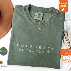 Könnte beinhalten: Ein moosgrünes T-Shirt mit den Worten "EMERGENCY DEPARTMENT" in Weiß aufgedruckt. Das Shirt hat einen Rundhalsausschnitt und kurze Ärmel. Eine goldene Halskette mit Anhänger ist auf dem Shirt drapiert. Das Shirt ist mit "Comfort Colors" gekennzeichnet.