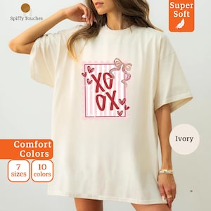 May include: An ivory-coloured t-shirt featuring a red and pink "XO XO" graphic within a pink-lined box. The shirt has short sleeves and a relaxed fit. The words "Super Soft" are in an orange box.