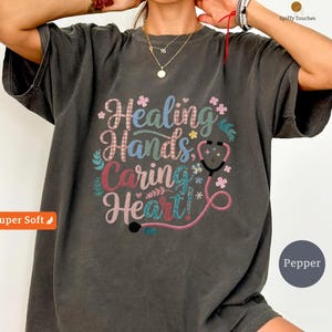 Peut inclure: T-shirt gris foncé avec l'inscription "Healing Hands, Caring Heart!" dans un motif floral coloré. Un graphique de stéthoscope est inclus. Le t-shirt a une texture douce et est étiqueté "Super Soft" et "Pepper".