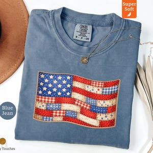 以下が含まれることがあります： 青いジーンズ色のTシャツ。縫い付けられたアメリカ国旗のデザインが特徴です。国旗は赤と白のストライプ、青い星、装飾的なボーダーが特徴です。ペンダント付きのシルバーネックレスがシャツの上にあります。シャツには「Super Soft」と表示されています。