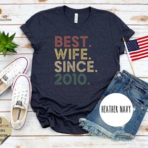Puede incluir: Una camiseta azul marino jaspeada con el texto "BEST. WIFE. SINCE. 2010." en una fuente de estilo vintage. El texto est&aacute; en tonos de rojo, naranja, amarillo y verde.