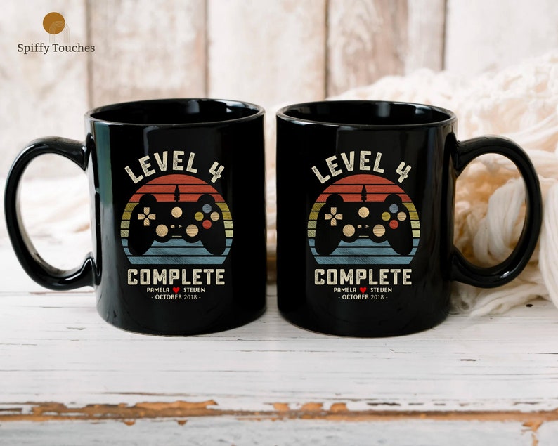 May include: Two black coffee mugs with a retro video game design. The mugs say "Level 4 Complete" with a date and names.