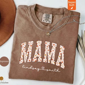 Pode incluir: Camiseta Comfort Colors cor espresso com a palavra "MAMA" em um design floral. O nome "lindsay smith" está escrito abaixo da palavra "MAMA". Um colar de prata está sobre a camiseta.