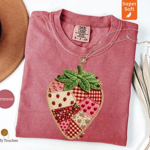 May include: Crimson-coloured t-shirt featuring an embroidered strawberry design. The strawberry is crafted from fabric patches with gingham, floral, and heart patterns. A silver necklace with a pendant is draped on the shirt.