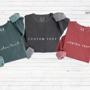 May include: Three long-sleeved sweatshirts in teal, charcoal gray, and brick red. Each sweatshirt has a crew neck and the text "custom text" printed on the front. The cuffs are a contrasting color.