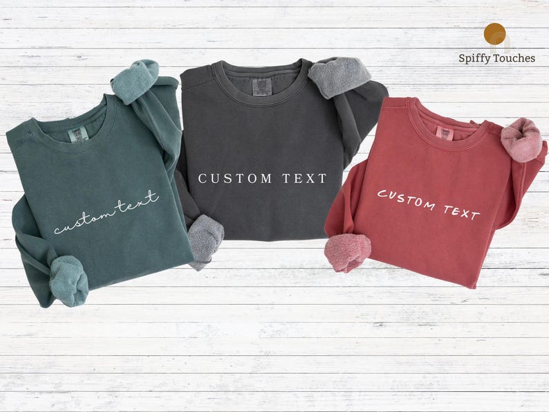 May include: Three long-sleeved sweatshirts in teal, charcoal gray, and brick red. Each sweatshirt has a crew neck and the text "custom text" printed on the front. The cuffs are a contrasting color.