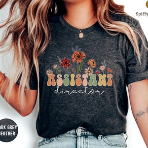 May include: Dark grey heather t-shirt with a floral design and the text "Assistant Director" in a retro font.