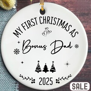 May include: White ceramic Christmas ornament with gold hanging loop. The ornament features the text "MY FIRST CHRISTMAS AS Bonus Dad" in black script, along with decorative snowflakes, hearts, and Christmas trees. The year 2025 is also displayed.