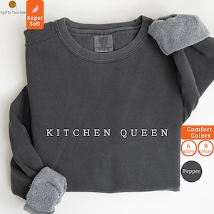 May include: A dark gray sweatshirt with the text "KITCHEN QUEEN" in white. The sleeves are cuffed with a gray, fuzzy material. The sweatshirt has a crew neck and is labeled "Comfort Colors".
