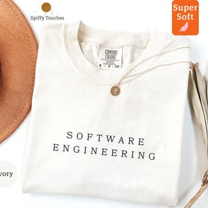 Può includere: T-shirt color avorio con la scritta "SOFTWARE ENGINEERING" stampata in nero. La maglietta ha girocollo e maniche corte. Una collana d'oro con un ciondolo poggia sulla maglietta. La maglietta è etichettata "Super Soft".