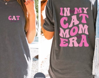 Könnte beinhalten: Ein dunkelgraues T-Shirt mit dem Text "IN MY CAT MOM ERA" in rosa und weißen Buchstaben mit Schmetterlingsakzenten. Der Text ist in einem Retro-Schriftart-Stil.
