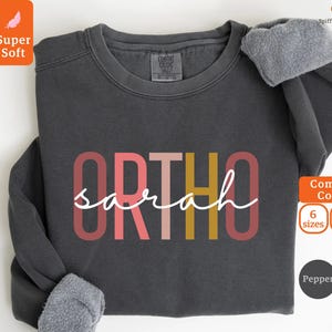 Könnte beinhalten: Ein dunkelgraues Sweatshirt mit dem Wort "ORTHO" in bunten Buchstaben und dem Namen "sarah" darunter in weißer Kursivschrift. Das Sweatshirt ist aus superweichem Material und in 6 Größen und 8 Farben erhältlich.