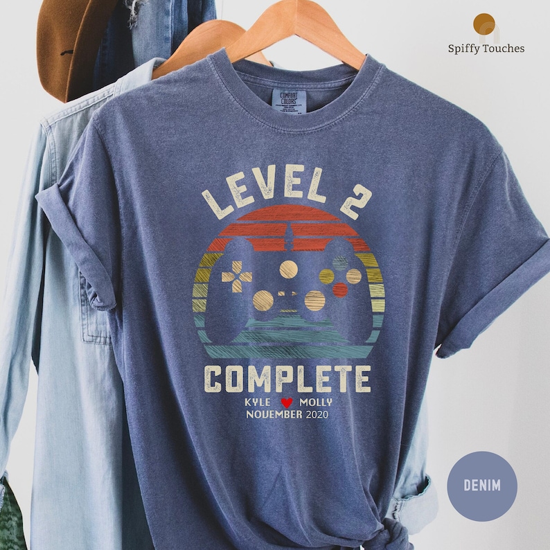 Op de afbeelding: Een blauw T-shirt met een vintage-stijl grafische afbeelding van een videogamecontroller en de tekst "Level 2 voltooid" met de namen "Kyle" en "Molly" en de datum "November 2020".