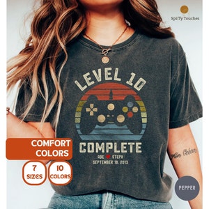 May include: A black t-shirt with a vintage-style graphic of a video game controller and the text "Level 10 Complete". The graphic is in shades of red, orange, yellow, blue, and green. The text "Abe Steph September 18, 2013" is printed below the graphic.
