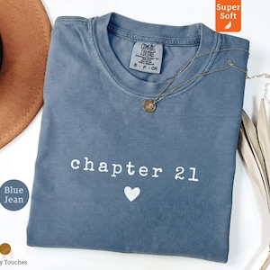 Puede incluir: Una camiseta de manga corta color azul jean con cuello redondo. La prenda presenta el texto blanco "chapter 21" impreso en el pecho, seguido de un pequeño icono de corazón blanco. La etiqueta interior indica "Comfort Colors" y la talla "S".