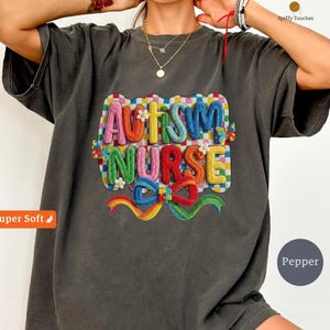 Puede incluir: Camiseta gris oscuro con las palabras bordadas "AUTISM NURSE" en colores vibrantes. El diseño incluye un lazo arcoíris y detalles florales. La camiseta tiene un corte holgado y está etiquetada como "Super Soft".