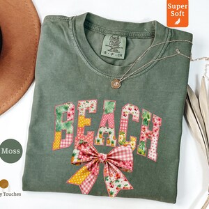 May include: A moss green t-shirt with the word "BEACH" in a patchwork design of floral and gingham patterns. A large pink bow with a gingham and floral pattern is below the word. A silver necklace with a pendant is on the shirt.