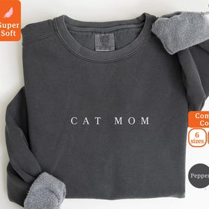 May include: Dark gray sweatshirt with the text "CAT MOM" in white. The sweatshirt has a crew neck and long sleeves with gray fleece cuffs. The image also includes text indicating "Super Soft" and "Comfort Colors" with options for sizes and colors.