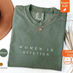 May include: A moss green t-shirt with the words "WOMEN IN AVIATION" printed in white. The shirt has a crew neck and short sleeves. A gold necklace with a pendant rests on the shirt. The shirt is labeled "Comfort Colors".