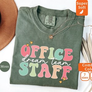Op de afbeelding: Een mosgroen T-shirt met de tekst "OFFICE dream team STAFF" in kleurrijke retro-lettertypen. Het shirt is gemaakt van een zachte, comfortabele stof en is verkrijgbaar in 7 maten en 10 kleuren.