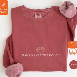 Può includere: Felpa color cremisi con la scritta "MAMA MAKES THE DOUGH" e una grafica di pane. La maglia a maniche lunghe ha uno scollo tondo e un interno morbido e strutturato. L'immagine mostra anche le parole "Super Soft" e "Comfort Colors".