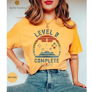 Op de afbeelding: Een mosterdgeel T-shirt met een retro videogamecontroller-afbeelding en de tekst "Level 2 Complete Kyle &hearts; Molly November 2020".