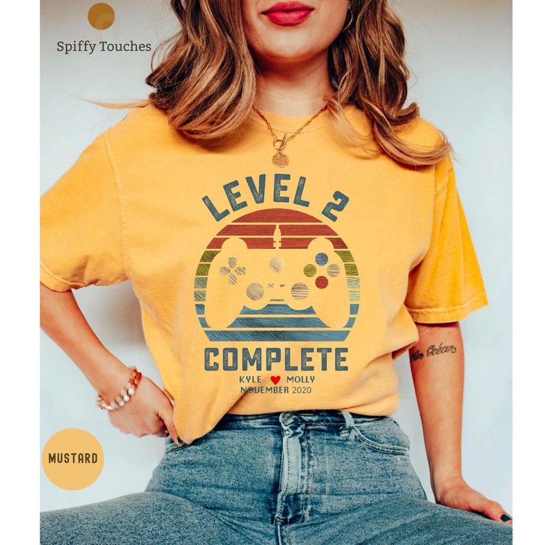 Op de afbeelding: Een mosterdgeel T-shirt met een retro videogamecontroller-afbeelding en de tekst "Level 2 Complete Kyle &hearts; Molly November 2020".