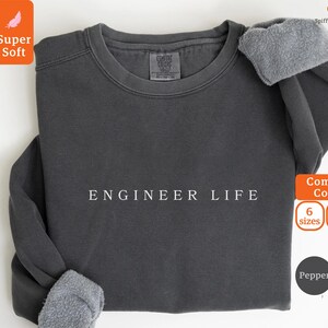 Könnte beinhalten: Ein dunkelgraues Sweatshirt mit dem weißen Text "ENGINEER LIFE". Das Sweatshirt hat einen Rundhalsausschnitt und lange Ärmel mit grauen Fleece-Bündchen. Das Bild enthält auch Text, der "Super Soft" und "Comfort Colors" anzeigt.