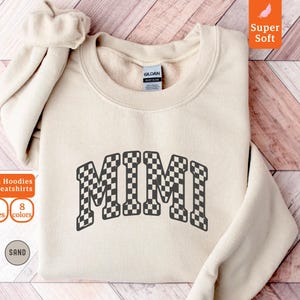 以下が含まれることがあります： 白と黒の市松模様で「MIMI」の文字が入ったサンドカラーのスウェットシャツ。スウェットシャツはクルーネックと長袖です。画像には「Gildan Hoodies & Sweatshirts」、「6サイズ」、「8色」、「Super Soft」という文字も含まれています。