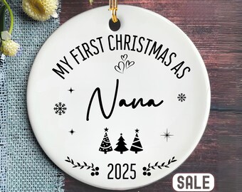 May include: White ceramic Christmas ornament with gold hanging loop. The ornament reads "MY FIRST CHRISTMAS AS Nana 2025" in black script and block letters, with decorative snowflakes, hearts, stars, and Christmas trees.