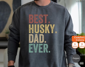 May include: Dark gray sweatshirt with the text "BEST. HUSKY. DAD. EVER." in red, yellow, and teal lettering. The sweatshirt has a crew neck and long sleeves. The image also shows the words "Comfort Colors", "6 sizes", "8 colors", and "Pepper".