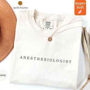 May include: Cream-coloured t-shirt with the word "ANESTHESIOLOGIST" printed in black. A gold necklace with a pendant rests on the shirt. The shirt is labelled "Comfort Colors" and "Super Soft".