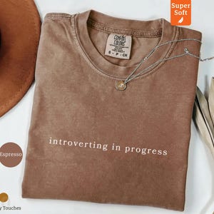 May include: Espresso-colored t-shirt with the phrase "introverting in progress" in white. The shirt has a crew neck and short sleeves. A silver necklace with a pendant is draped on the shirt. The shirt is labeled "Super Soft".