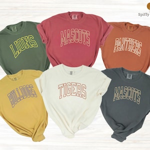 May include: Six short-sleeved t-shirts in various colors, each with a different team name in a collegiate font. Colors include olive green, red, orange, yellow, white, and gray. The text on the shirts reads "LIONS", "MASCOTS", "PANTHERS", "BULLDOGS", "TIGERS", and "MASCOTS".