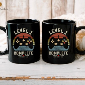 May include: Two black coffee mugs with a retro-style video game controller graphic and the text "Level 1 Complete". The mugs are personalized with the names "Pamela" and "Steven" and the date "October 19, 2021".