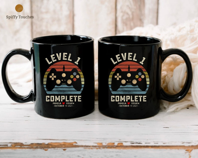 May include: Two black coffee mugs with a retro-style video game controller graphic and the text "Level 1 Complete". The mugs are personalized with the names "Pamela" and "Steven" and the date "October 19, 2021".