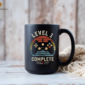 May include: Black ceramic coffee mug with a retro-style design featuring a video game controller and the text "Level 1 Complete". The mug also includes the names "Pamela" and "Steven" and the date "October 19, 2021".