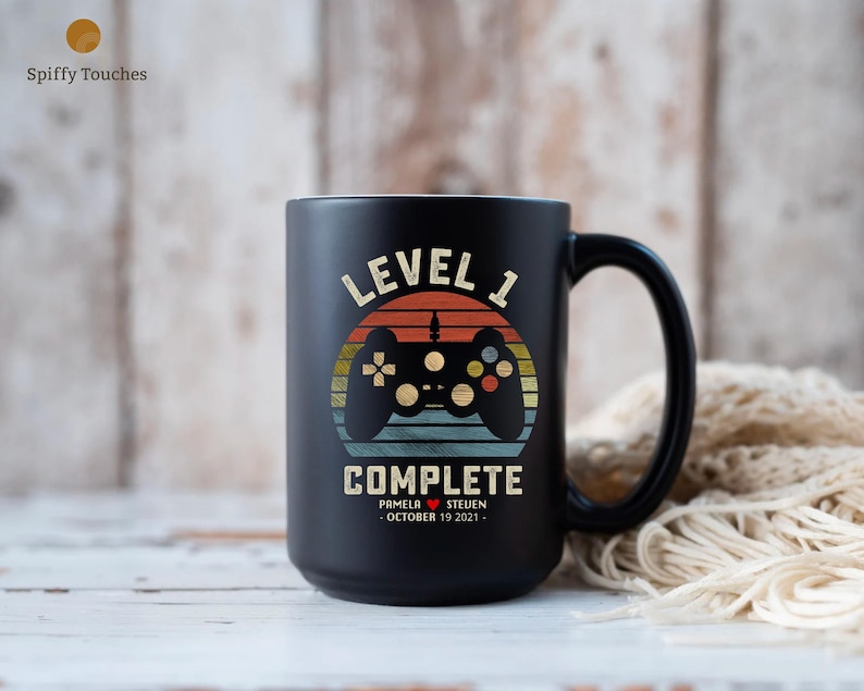 May include: Black ceramic coffee mug with a retro-style design featuring a video game controller and the text "Level 1 Complete". The mug also includes the names "Pamela" and "Steven" and the date "October 19, 2021".