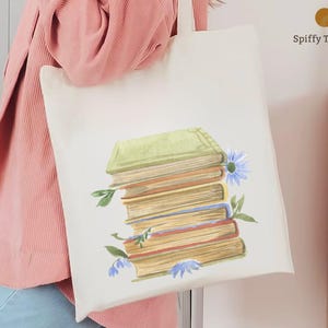 Op de afbeelding: Crèmekleurige draagtas met een aquarelillustratie van een stapel boeken. De boeken hebben groene, blauwe en bruine covers, met blauwe bloemen en groene bladeren. De tekst "Spiffy Touches" staat in de rechterbovenhoek.