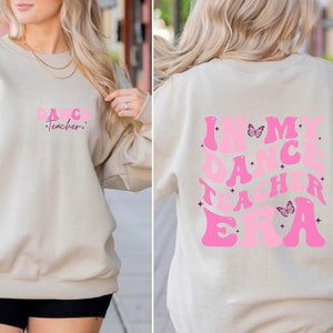 May include: A light beige sweatshirt with the text "Dance Teacher" in pink letters on the front and "In My Dance Teacher Era" in pink letters on the back. The back design also includes two pink butterflies.