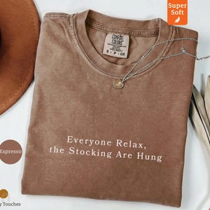 May include: Espresso-colored Comfort Colors t-shirt with the text "Everyone Relax, the Stocking Are Hung" in white. A silver necklace with a pendant is draped on the shirt. The shirt is displayed on a white surface.