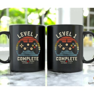 May include: Two black coffee mugs with a retro video game controller graphic and the text "Level 1 Complete" with a date and names.