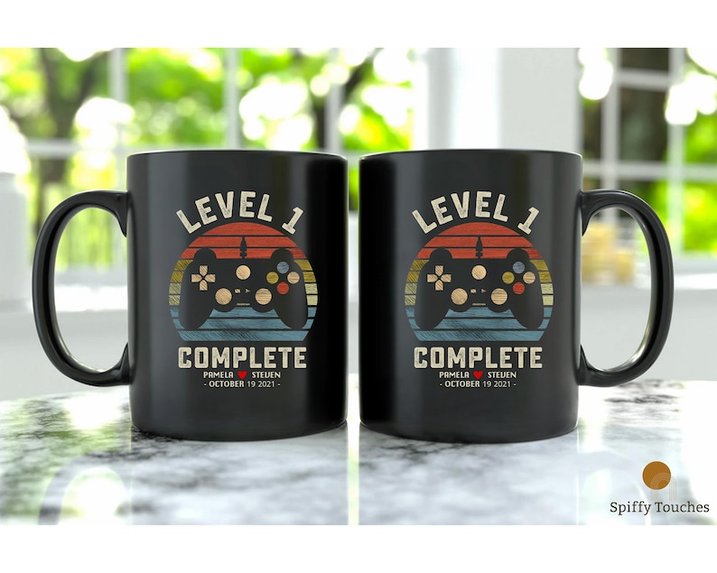May include: Two black coffee mugs with a retro video game controller graphic and the text "Level 1 Complete" with a date and names.