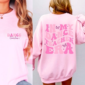 May include: Pink sweatshirt with the text "In My Dance Teacher Era" in pink and white letters with butterfly graphics. The sweatshirt has a crew neck and long sleeves.
