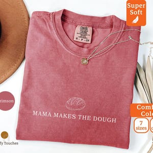 Może przedstawiać: Karmazynowa koszulka z napisem "MAMA MAKES THE DOUGH" i grafiką chleba. Koszulka ma okrągły dekolt i krótkie rękawy. Złoty naszyjnik z wisiorkiem znajduje się na koszulce. Koszulka jest z Comfort Colors i jest bardzo miękka.