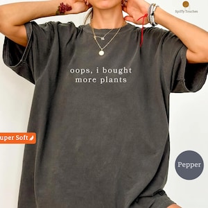 Puede incluir: Camiseta gris oscuro con el texto blanco "oops, i bought more plants". La camiseta tiene un corte relajado y está etiquetada como "Super Soft" y "Pepper". Una camiseta informal y cómoda.
