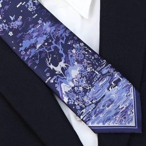 May include: A navy blue necktie featuring a detailed illustration of a deer, trees, and water in shades of blue and purple. The tie is made of a smooth fabric and is worn with a white collared shirt and a dark suit jacket.